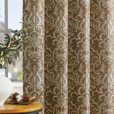 a vase with an olive plant in it next to a window covered in brown and white damask curtains