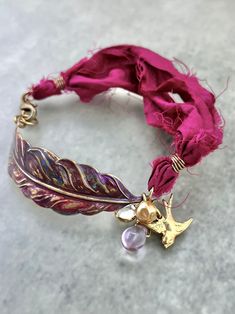 WHAT IT IS: Handmade bohemian-style bracelet featuring upcycled magenta sari silk, a large hand-enameled and hand-formed brass feather, a brass bird charm, a small vintage Swarovski channel-set crystal, and vintage beads. THE SPECS: WHY IT'S SPECIAL: The brass feather used in this bracelet was enameled by hand in shades of magenta, purple, burgundy, and blue. The ribbon is upcycled from torn and dyed silk that was leftover from the sari manufacturing process. Bohemian Purple Jewelry For Festive Occasions, Purple Bracelets For Festivals And Gifts, Sari Ribbon Jewelry, Silk Ribbon Jewelry, Sari Silk Jewelry, Brass Bird, Sari Ribbon, Purple Burgundy, Artisan Jewelry Handmade