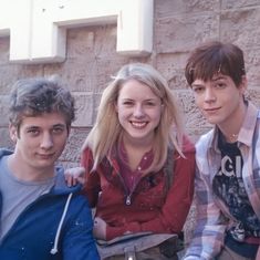 three young people sitting next to each other on the ground near a stone wall and building