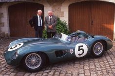 two men standing next to a green race car on a cobblestone driveway in front of garage doors