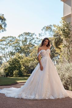 a woman in a wedding dress standing on the ground near some trees and bushes with her hand on her hip