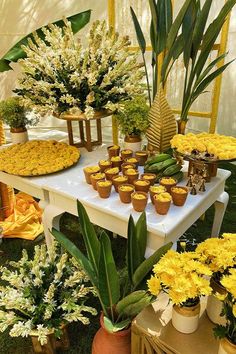 a table topped with lots of yellow flowers and cupcakes next to potted plants