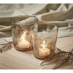 two glass candles sitting on top of a wooden table next to pine cones and branches