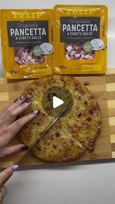 a person cutting up a pizza on top of a wooden cutting board next to two packages of pancetta