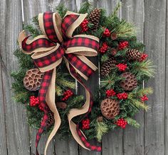 a christmas wreath with pine cones and berries hanging on a wooden fence next to evergreens