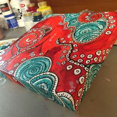 a red and blue pillow sitting on top of a table next to bottles of paint