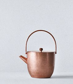 a copper tea pot with a handle on a white surface, next to a gray wall