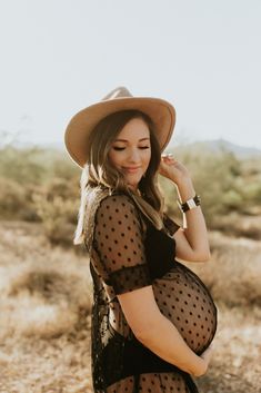 a pregnant woman in a black dress and hat poses for the camera with her hands on her hips