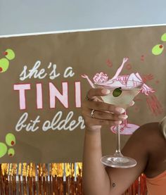 a woman holding up a martini glass in front of a sign