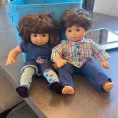 two dolls sitting next to each other on top of a wooden table in front of a blue bin