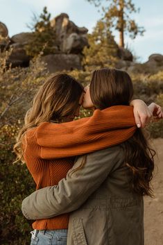 two women hugging each other while standing in front of some rocks and trees on a sunny day