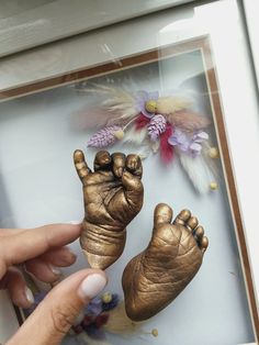 a person is holding a gold baby foot in front of a frame with flowers and feathers on it