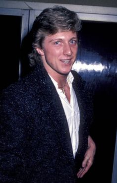 a man in a black jacket and white shirt standing next to a door with his hands in his pockets