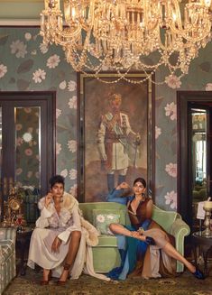 two women sitting on couches in front of a wall with paintings and chandeliers