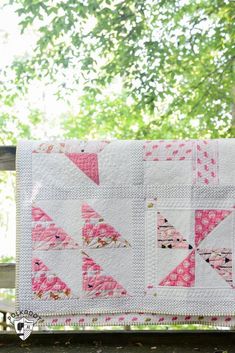 a pink and white quilt hanging on a wooden fence with trees in the back ground