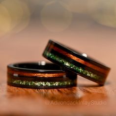 two wedding bands with green and gold inlays on them sitting on a wooden surface