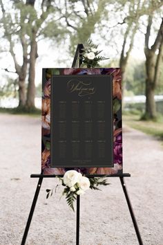 a sign with flowers on it sitting in the middle of a dirt road next to trees