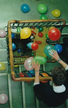 two people standing in front of a mirror with balloons on the wall behind them and one person reaching up to grab something