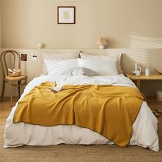 a bed with white and yellow sheets in a bedroom next to a table, chair and lamp