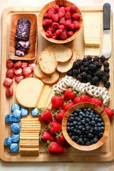 a wooden tray topped with fruit and crackers next to bowls of cheese, strawberries and blueberries