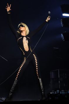 lady in black outfit performing on stage with microphone and hands raised up to the sky