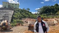 a man standing in the dirt next to a bulldozer and a building on top of a hill