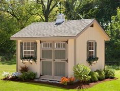 a small shed with two windows and flowers in the front yard, next to some trees