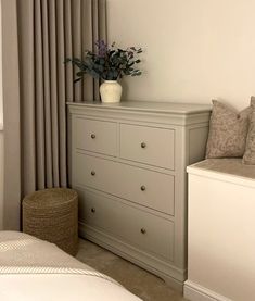 a white vase with flowers on top of a dresser next to a bed and window