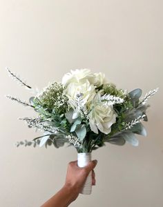 a person holding a bouquet of white flowers and greenery in front of a wall