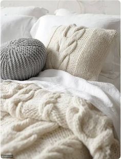 a white bed topped with two knitted pillows next to a pillow on top of a blanket