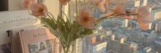 a vase filled with pink flowers next to a book on a window sill in front of a cityscape