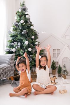 two young children sitting in front of a christmas tree with their hands up to the air