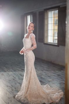 a woman in a long sleeved wedding dress standing on a wooden floor with her arms behind her back