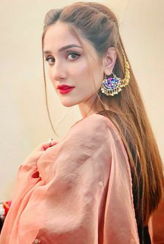 a woman with long hair wearing earrings and a pink top standing in front of a white wall