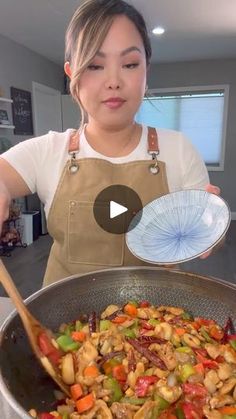 a woman in an apron holding a pan filled with stir fry vegetables and meats