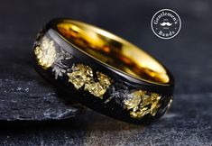 a black and gold wedding band with flowers inlayed to the side, sitting on top of a rock