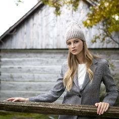 a woman leaning on a wooden fence wearing a gray jacket and white shirt with a knitted hat