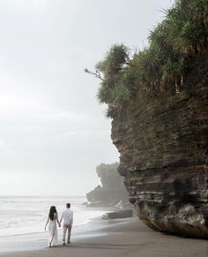 a man and woman walking on the beach next to an ocean cliff with grass growing out of it