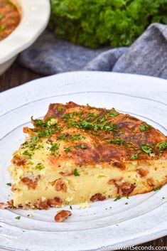 a piece of quiche on a white plate next to a bowl of greens and bread