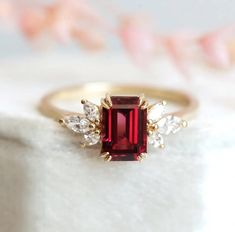 Made to orderThis exquisite ring showcases a mesmerizing emerald-cut red garnet that exudes a deep, rich hue reminiscent of passion and vitality. The intricate cluster of Moissanite marquise cut stones on either side enhances the ring's brilliance, creating a stunning play of light. Meticulously crafted from solid gold, this ring promises enduring shine and exceptional quality. Whether chosen for its striking beauty or as a symbol of love and intensity, our Red Garnet Ring epitomizes elegance an Red Things, Garnet Color, Dream Rings, Garnet And Diamond Ring, Garnet Engagement Ring, Red Garnet Ring, Floral Engagement Ring, Alexandrite Ring, Ring Ideas