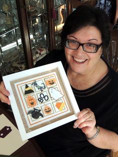 a woman sitting at a table holding up a framed halloween art piece in front of her