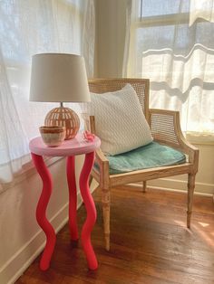 a pink table sitting next to a window with a lamp on it and a pillow