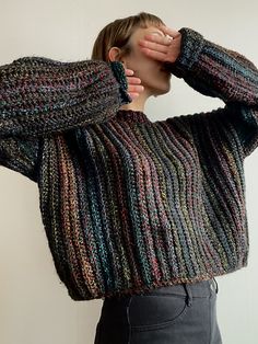 a woman wearing a multicolored sweater covering her face with one hand while standing in front of a white wall