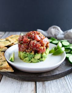 a white plate topped with an avocado covered in toppings next to sliced cucumbers and crackers