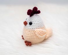 a small crocheted chicken sitting on top of a white fur covered floor next to a red flower