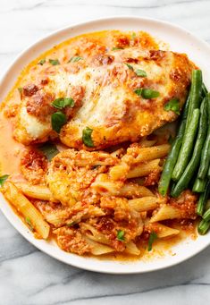 a white plate topped with pasta and meat covered in sauce next to green beans on a marble surface