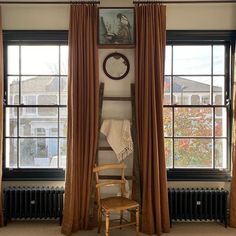 a chair sitting in front of two windows with drapes on them and a clock above the window