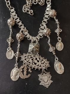 "A bib style assemblage of Victorian silver and religious medals. The central pendant is a Victorian brooch with chased and pierced floral and vine motif. Suspended from the brooch is a Victorian silver stick pin charm monogrammed letter \"C\". The other charm is a hallmarked sterling Victorian watch fob with the initials \"GW\" inside a heart. On the back is in engraved \"AL to GW \" and dated \"24/3/04\" for March 24, 1904. I've used a vintage silver plated Art Deco style bracelet link to attach it to the silver plated book chain.  The accompanying pendants are Bali sterling beads, faceted pyrite and religious Catholic medals from the 1930s or older featuring Our Lady of Lourdes. I believe they are plated silver. Finishes with larger links and hook so length is adjustable." Vintage Metal Jewelry With Intricate Design, Ornate Metal Jewelry Vintage Collection, Ornate Metal Jewelry For Vintage Collection, Traditional Vintage Charm Pendant Jewelry, Vintage Silver Pendant Jewelry, Traditional Pendant Jewelry With Vintage Charm, Silver Medallion Necklace With Charms, Antique Cross Necklaces For Collectibles, Antique Cross Necklace For Jewelry Making