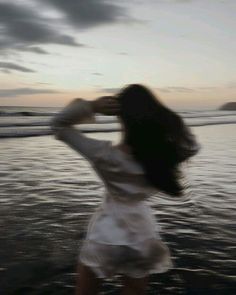 a woman standing on top of a beach next to the ocean at sunset with her hair blowing in the wind