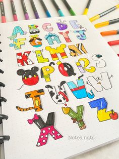 an open notebook with letters and numbers drawn on it next to colored markers, pencils and pens